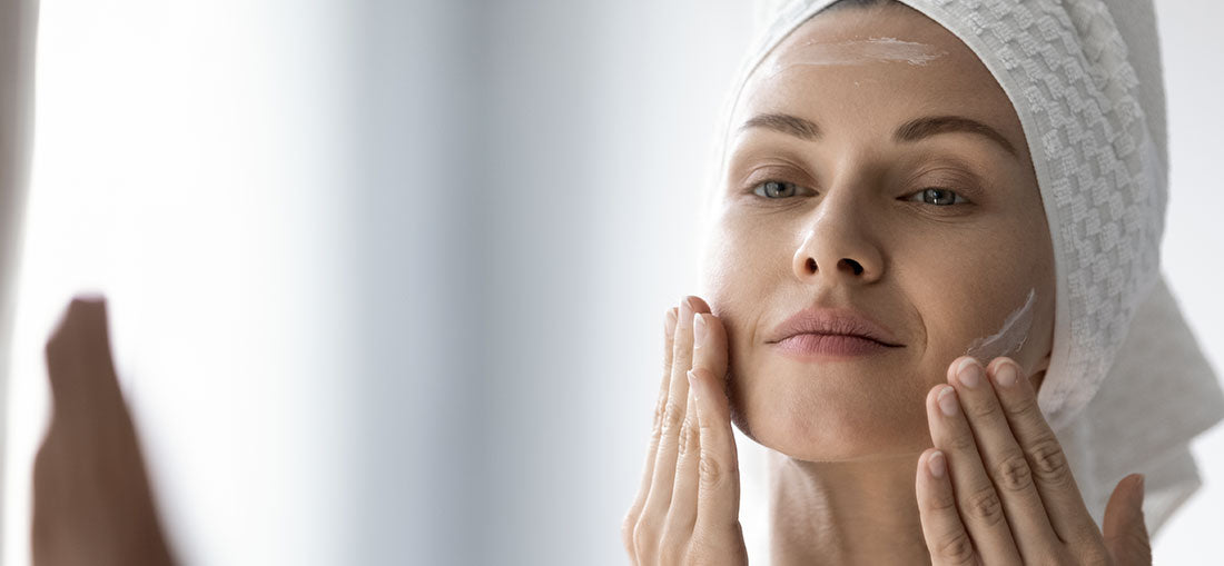woman wearing towel on her head applying moisturizer to cheeks in the mirror